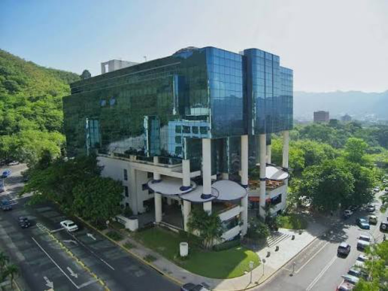 Vista aérea real del edificio del Instituto Docente de Urología en La Viña, Valencia: la torre de vidrio azul elevada sobre columnas blancas, rodeada de verde con la montaña detrás