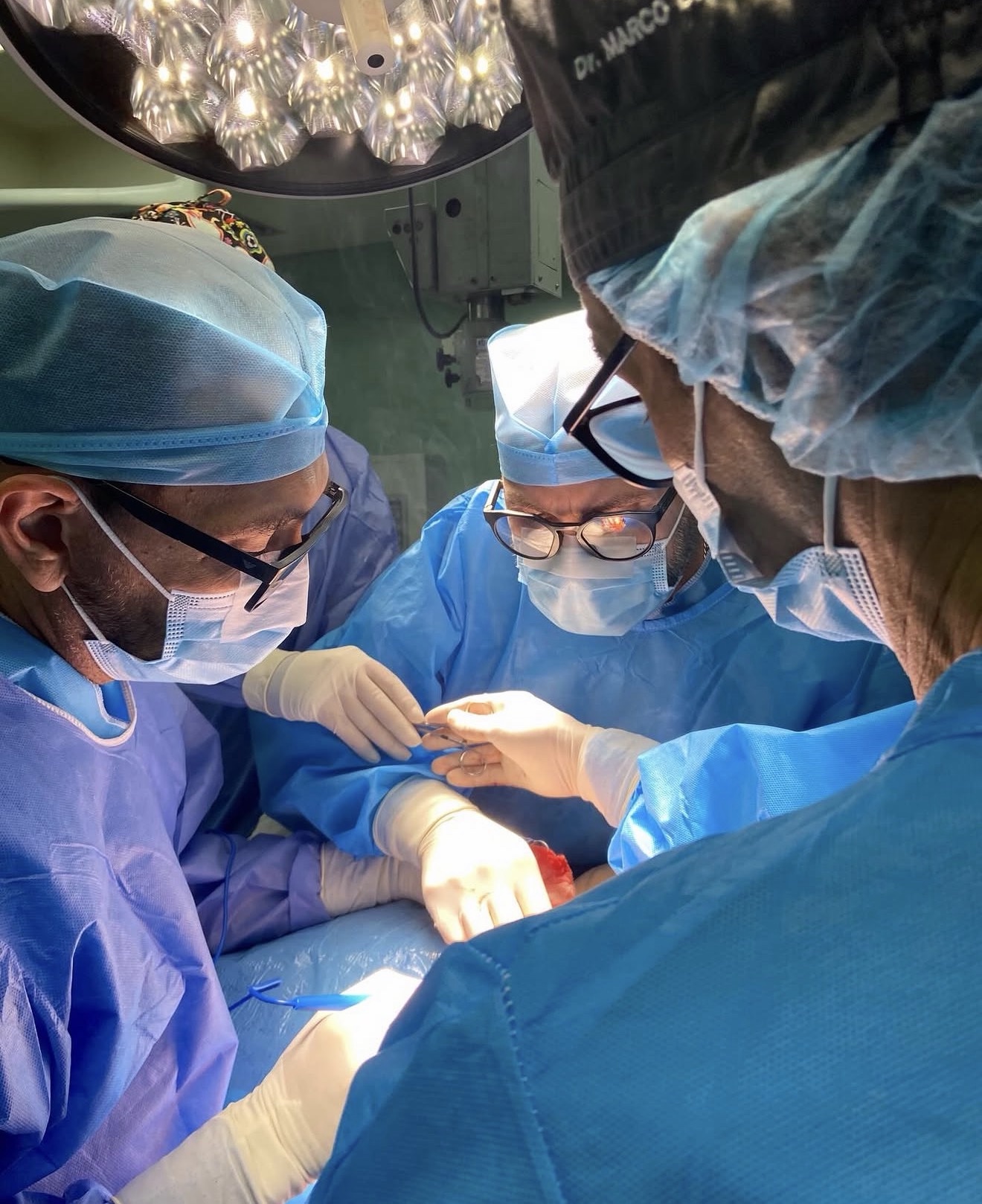 Equipo médico del IDU operando en quirófano bajo la lámpara quirúrgica — se lee «Dr. Marco» en el gorro de uno de los cirujanos — fotografía real del instituto